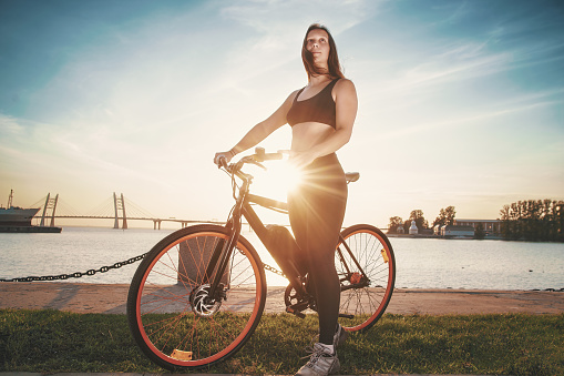 Athletic girl staying with electric bike against urban seascape. Outdoor portrait at sunset