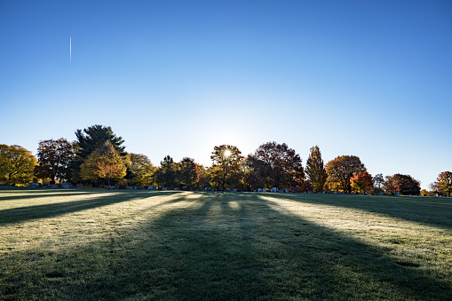 A jet airliner leaves a vertical contrail vapor trail rising above a rural cemetery with the lens flare sun rising through the trees and throwing long shadows above the graves and tombstones and beyond across the quiet morning dew-drenched meadow.