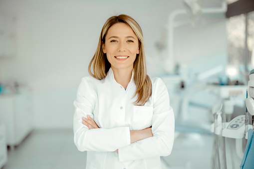 Young blonde woman dentist posing at modern dental cabinet, with arms crossed and cheerfully smiling at camera, copy space. Pretty stomatologist at dental clinic