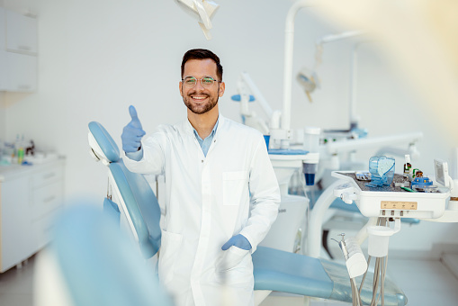 Male dentist in doctors white lab coat posing with thumb up in modern dental office
