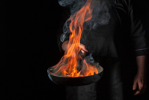 Chef cooking food in pan with fire flame on black background. Restaurant and hotel service concept. Free advertising space