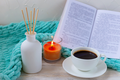 A cup of black coffee, aroma reed diffuser, a burning candle, an open book and a turquoise blanket. The concept of peace and relaxation. Home comfort
