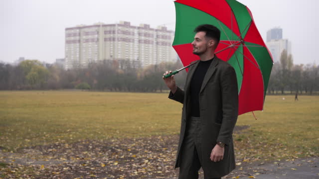 Handsome young man standing with green and red umbrella on cloudy misty day outdoors looking around. Portrait of confident positive Caucasian guy enjoying overcast day. Lifestyle and weather.