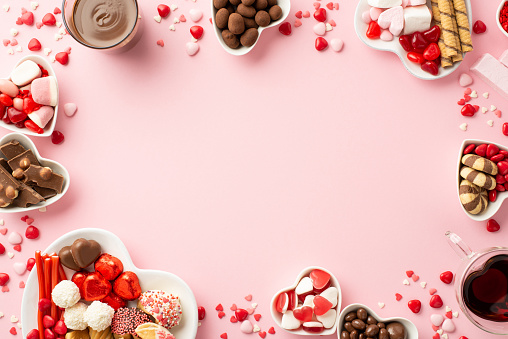 Valentine's Day concept. Top view photo of heart shaped saucers with sweets candies and glasses with beverage on isolated pastel pink background with copyspace in the middle