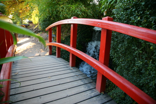 a red, wooden brigde in a japanese garden, leading over a creek with a little waterfall