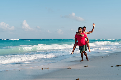 homosexual couple of two men playing piggybacking on the shore of cancun beach, vacation concept