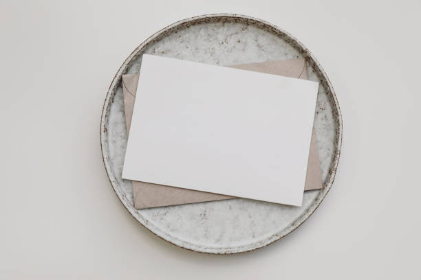 istock Neutral greeting card mockup. Blank paper invitation, craft envelope on ceramic plate isolated on white table background. Empty copy space. Flat lay, top view, nobody. Elegant stationery template. 1443378744