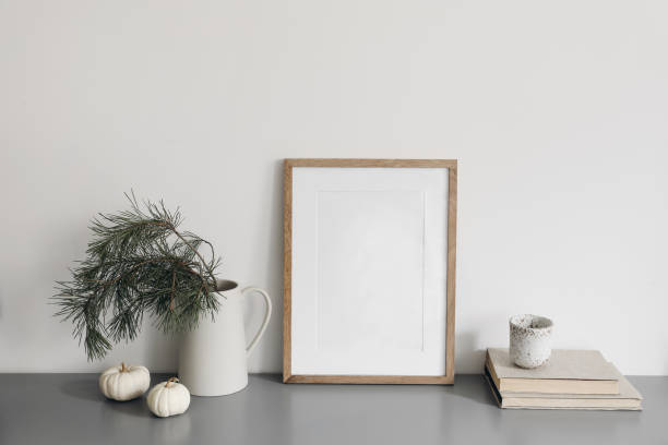 istock Christmas interior decor, still life. Blank wooden picture frame mockup. Vase with pine tree branches, cup of coffee and old books on grey desk. Winter home office. White little pumkins on table. 1443378749