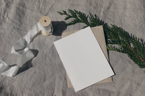 istock Winter wedding mockup. Empty greeting card, invitation mock up template with silk ribbon, cypress tree branch in sunlight. Craft envelope on linen tablecloth. Christmas still life. Flat lay, top view. 1443378810