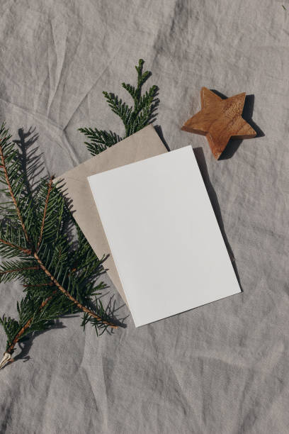 Natural Christmas stationery still life. Blank greeting card, invitation mock up with wooden star ornament and fir, cypress tree branch in sunlight. Craft envelope, linen tablecloth. Vertical top view stock photo