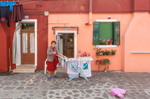 Burano, Italy - April 10, 2007: elderly woman puts her washing on a drying rack in front of her colorful house in Burano, Venice, Italy.