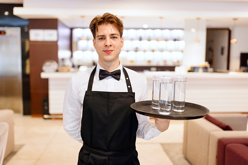 Male waiter carrying tray with glasses of water in hotel cafe.