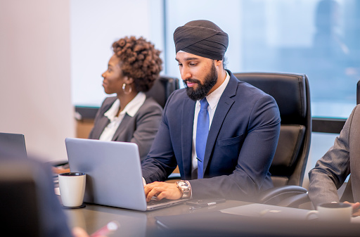 A group of mature adults sit at a boardroom table as they listen in attentively at a corporate business meeting.  They are each dressed professionally and have paper out in front of them as they take notes.