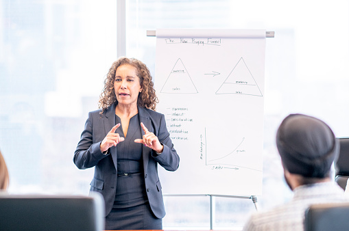A woman stands at the head of a boardroom table as she leads her team in a meeting.  She is dressed professionally and is standing on front of an easel as she uses it to teach her strategy.