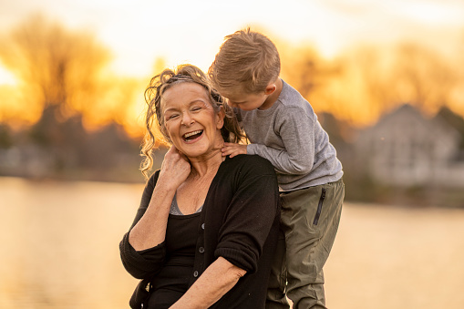 A senior woman and her Grandson pose together outside for a portrait.  They are both dressed casually and are embracing as they spend quality time together.