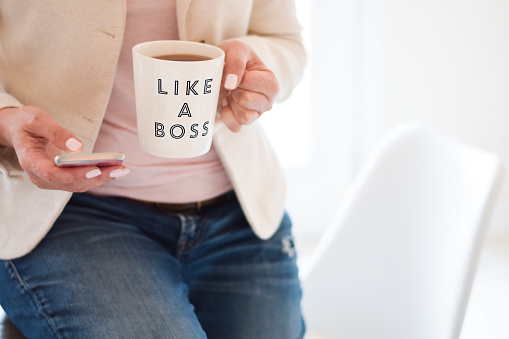 Midsection of business woman leaning on a desk using mobile phone and holding a cup that says Like a Boss with copy space