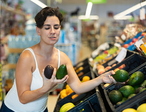 Woman choosing fresh avocado in supermarket in fruits and vegetables department.