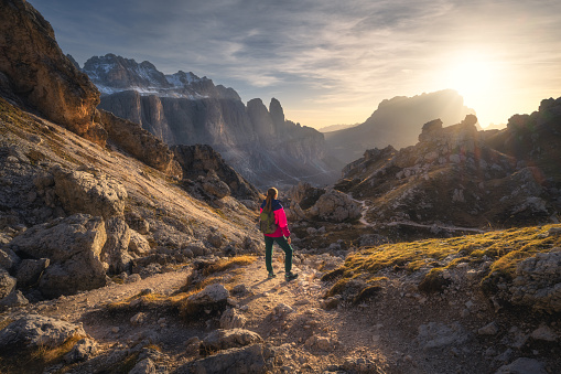 Girl with backpack on the trail in mountains at sunset in autumn. Beautiful landscape with young woman, high rocks, path, stones, orange grass, sky with clouds in fall in Dolomites, Italy. Adventure