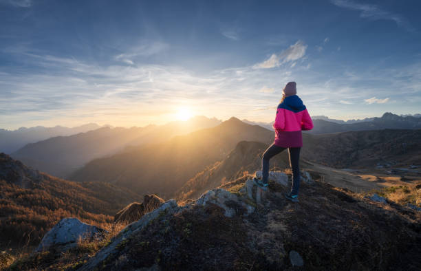 Young woman on mountain peak and beautiful mountains in haze at colorful sunset in autumn. Dolomites, Italy. Sporty girl, mountain ridges in fog, orange grass, trees, golden sun in fall. Hiking stock photo
