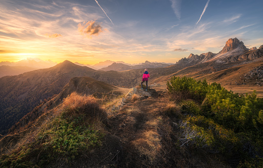 Young woman on the stone on mountain peak and beautiful mountains at colorful sunset in autumn. Dolomites, Italy. Sporty girl, mountain ridges, orange grass, green trees, golden sun in fall. Hiking