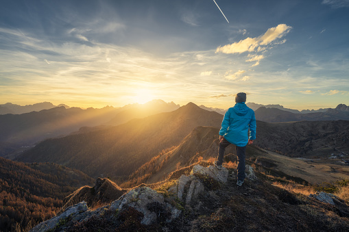 Hombre en piedra en la colina y hermosas montañas en bruma en la colorida puesta de sol en otoño. Dolomitas, Italia. Chico deportista, crestas montañosas en niebla, hierba naranja y árboles, cielo azul con sol en otoño. Excursionismo photo