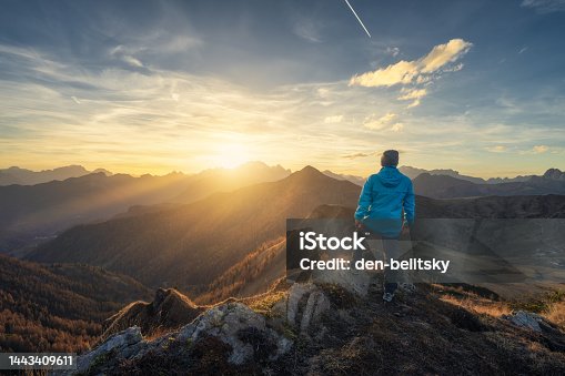 istock Man on stone on the hill and beautiful mountains in haze at colorful sunset in autumn. Dolomites, Italy. Sporty guy, mountain ridges in fog, orange grass and trees, blue sky with sun in fall. Hiking 1443409611