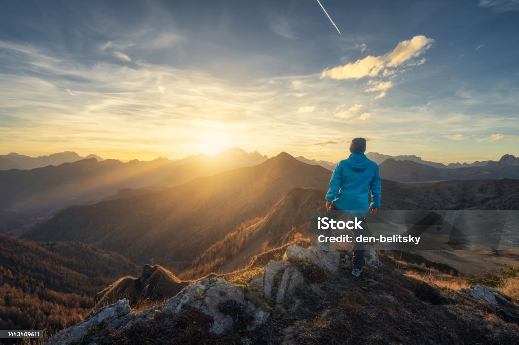 Man on stone on the hill and beautiful mountains in haze at colorful sunset in autumn. Dolomites, Italy. Sporty guy, mountain ridges in fog, orange grass and trees, blue sky with sun in fall. Hiking Mountain Stock Photo