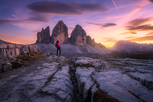 Girl on the mountain peak and high rocks at colorful sunset in autumn. Tre Cime, Dolomites, Italy. Beautiful landscape with young woman on trail, cliffs, purple sky with pink clouds in fall. Hiking