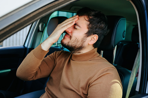A young man guy sits in a car holding his hand on his head which hurts. The driver behind the wheel stopped to catch his breath. He probably has a headache.