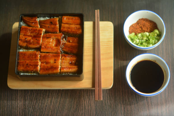 A beautifully plated Unagi Kabayaki (grilled Japanese eel) on a traditional bento box, served with steamed rice. The indoor setting offers a warm, inviting atmosphere with soft lighting, perfect for showcasing this delectable Japanese dish. Ideal for food bloggers, culinary websites, or anyone highlighting Japanese cuisine in an elegant and comforting environment.