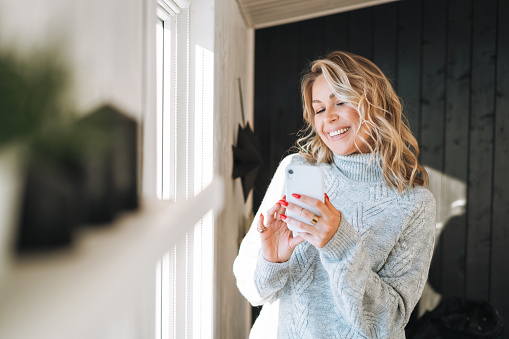 Young woman with blonde curly hair in grey sweater using mobile phone in hands near window at house