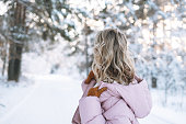 Young woman with blonde hair in winter clothes hands near face against background of snowy winter forest, view from back
