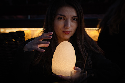 A beautiful young brunette girl with long hair holds a night lamp burning with warm yellow light in her palms and looks into the camera while sitting in a night room with a blurred background, close-up from the side, close-up from the side.