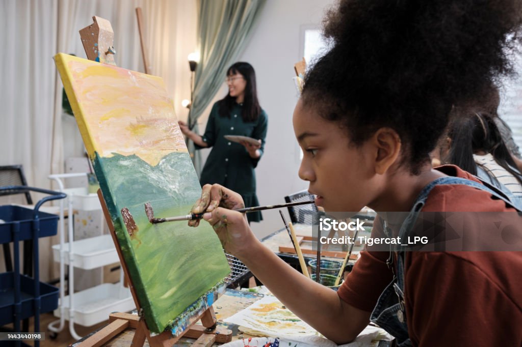 A girl concentrates on acrylic color painting on canvas in an art classroom. African American girl concentrates on acrylic color picture painting on canvas with students group in art classroom, creative learning with talents and skills in elementary school studio education. Art Stock Photo