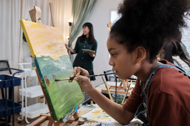 A girl concentrates on acrylic color painting on canvas in an art classroom. African American girl concentrates on acrylic color picture painting on canvas with students group in art classroom, creative learning with talents and skills in elementary school studio education. workshop art studio art paint stock pictures, royalty-free photos & images