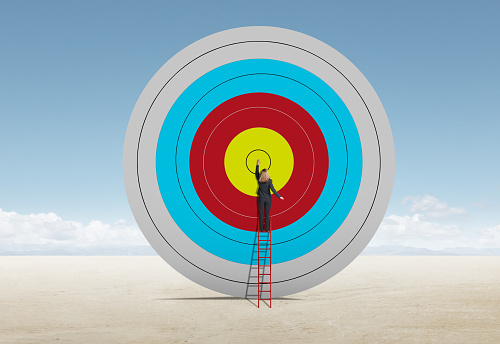 A woman stands on a tall red ladder as she reaches up and places her hand on the bull's eye of a target.