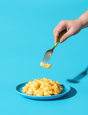 Plate with scrambled eggs in bright light minimalist on a blue table. Woman's hand taking scrambled eggs with a fork from a blue plate.