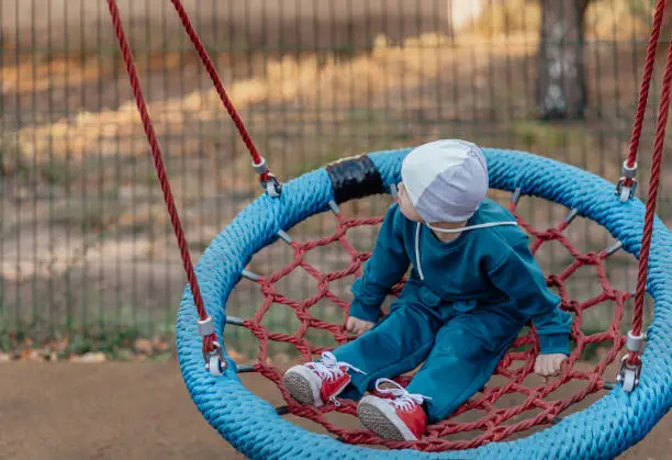 Photo of Cute Little boy with down syndrome in a funny hat walks in the playground, swinging on a swing