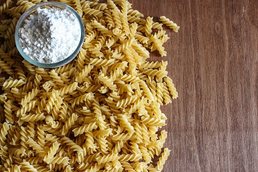 A top view of a pile of raw fusilli pasta and a bowl of flour on a wooden board