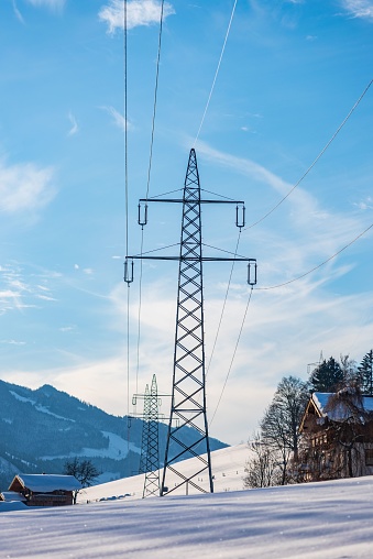 High voltage power pole with an overhead line. Winter scenery around, snow, trees and blue sky. Mountain landscape in the Alps.