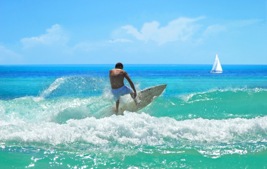 Surfer catching wave on gogeous turquoise sea with sailboat in the distance