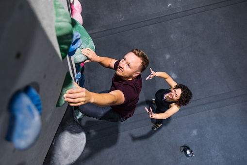 Climbing instructor assisting a man student during the climbing in a climbing wall. Man on the bouldering training with female instructor at sport climbing gym.