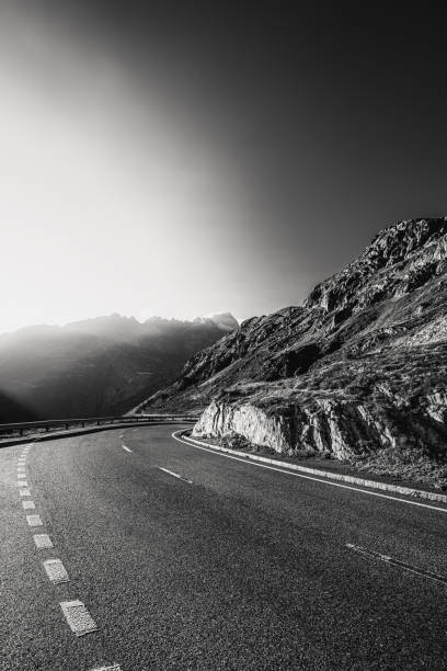 picture in black and white, view over the road going into a bend, on the right a rocky mountain face, sky and the early morning sun, which is still low, near the Furkapass, canton of Valais on the border with canton of Bern, switzerland