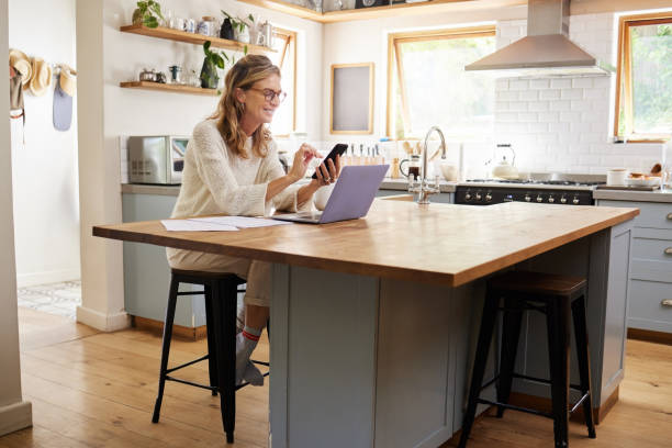 istock Woman, phone and kitchen smile for social media, email or text while work online. Mature, lady and smartphone with laptop for communication in remote job on web, laughing at meme or joke on cellphone 1443474906