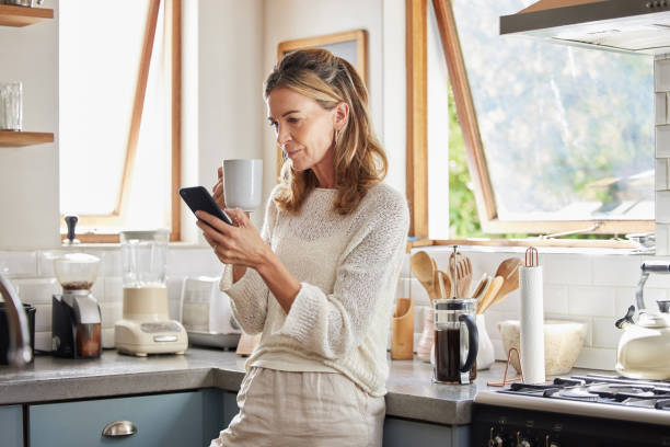 café, telefone e mulher idosa na cozinha navegando nas mídias sociais, mensagens de texto ou aplicativo on-line em casa. relaxe, chá e lendo notícias ou navegação na web on-line móvel com mulheres do canadá em smartphone 5g - bebida branca - fotografias e filmes do acervo