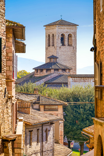 A glimpse of a harmonious stone alley in the historic center of the medieval town of Assisi in Umbria, central Italy, with the bell tower of the Chiesa dell'Abazia di San Pietro (St Peter's Abbey Church), built in Romanesque and Gothic style in the 10th century, in background. Famous for being the city of San Francesco and Santa Chiara (St Francis and St Clare), Assisi is recognized in the world as a place of spirituality and peace between all peoples and different confessions. The Umbria region, considered the green lung of Italy for its wooded mountains, is characterized by a perfect integration between nature and the presence of man, in a context of environmental sustainability and healthy life. In addition to its immense artistic and historical heritage, Umbria is famous for its food and wine production and for the high quality of the olive oil produced in these lands. Since 2000 the Franciscan sites of Assisi have been declared a World Heritage Site by UNESCO. Image in high definition format.