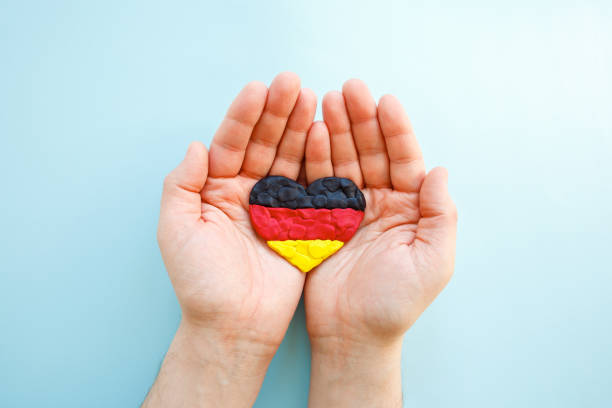 флаг германии. форма сердца из черного, красного и желтого пластилина моделирует глину в мужских руках на синем фоне. концепция немецкого ф� - german culture flash стоковые фото и изображения