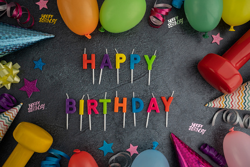 Happy Birthday cake candles letters with gym dumbbells, colorful balloons, cone party hats, bows, streamers and ribbons. Exercise equipment as a gift idea. Healthy fitness lifestyle flat lay concept.