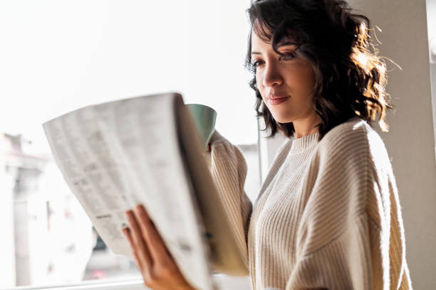 istock Young woman drinking cup of coffee while reading newspaper 1443493537