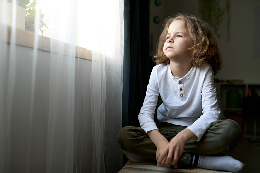 Pensive little boy sitting in his room and looking at window with thoughtful sight
